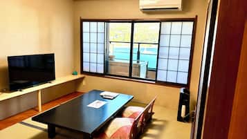 Una habitación con suelo de tatami, una mesa baja, un televisor sobre un mueble, una puerta corredera con vistas y un equipo de aire acondicionado.