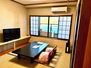 Una habitación con suelo de tatami, una mesa baja, un televisor sobre un mueble, una puerta corredera con vistas y un equipo de aire acondicionado.