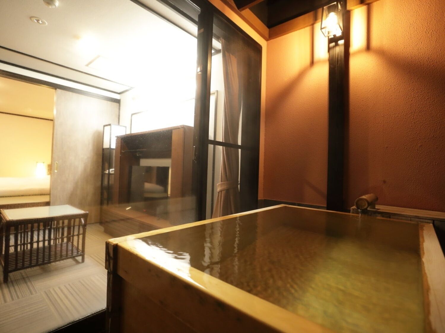 A hot spring bath area with a wooden bench, a glass partition, and a wall-mounted light fixture.
