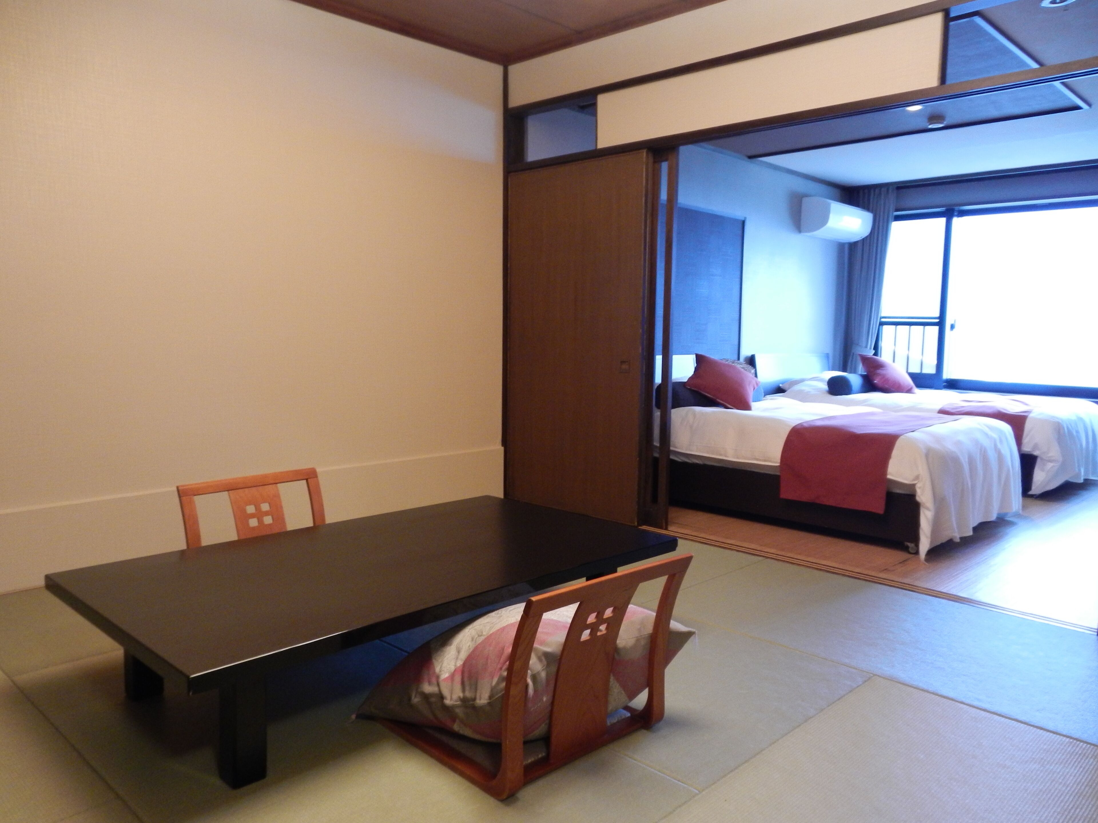 A room with a dark wooden table, a chair, and a bed with red bedding.