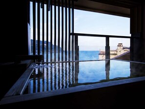 A room with a large window, a wooden bench, and a view of the sea and buildings. - Inatori Akao Hotel (Higashiizu)