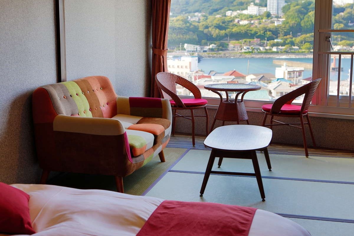 A hotel room with a bed, a sofa, a small table, and two chairs. The room has a view of a body of water and buildings in the distance.