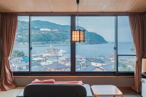 A room with a large window, a bed, a small table, and a hanging lamp.