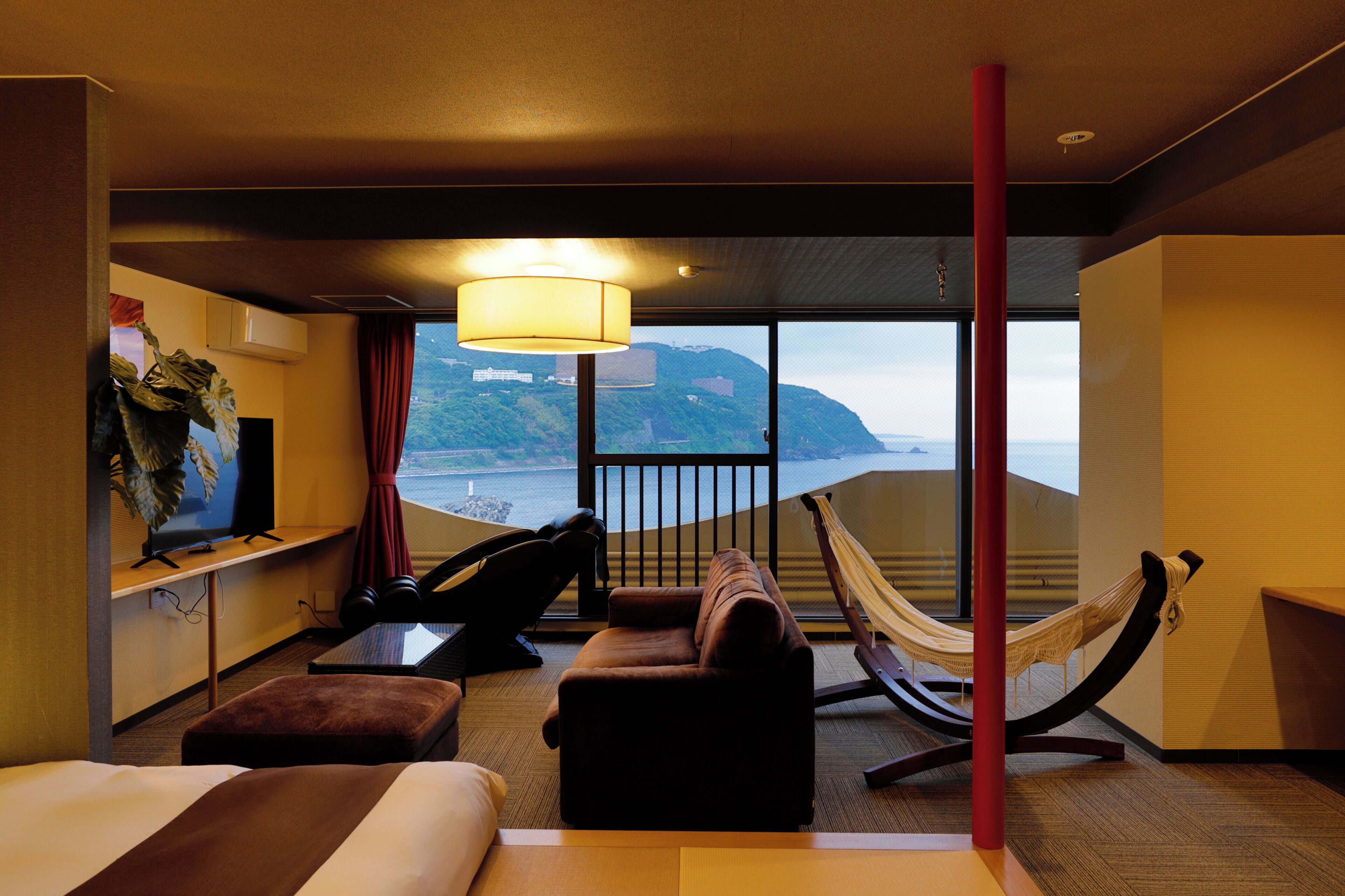 A hotel room with a bed, a desk, a chair, a sofa, a hammock, and a large window with a view of the sea and hills.