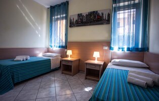 Habitación de hotel con dos camas individuales, cada una con colcha azul a rayas y almohadas blancas. Hay dos mesitas de noche con lámparas, y un cuadro de un paisaje urbano en la pared.