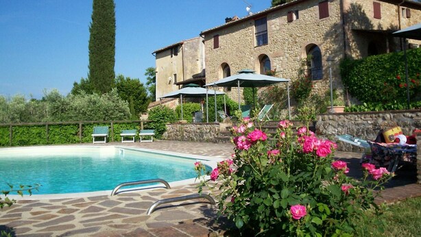 A pool area with a stone deck, lounge chairs, and a building with arched windows. - Podere Il Caggio Appartements (San Gimignano)