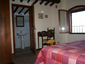 A bedroom with a bed, a wooden chair, a window, and a bathroom with a sink. - Podere Il Caggio Appartements (San Gimignano)
