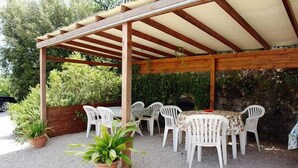 Een overdekt terras buiten, met een houten pergola, witte plastic stoelen en een tafel.