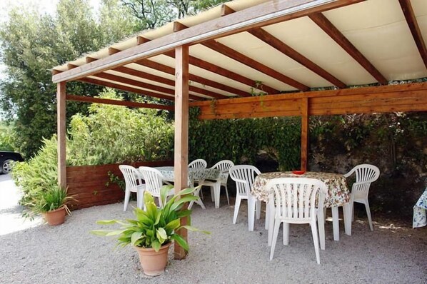 An outdoor covered patio with a wooden pergola, white plastic chairs, and a table. - Podere Il Caggio Appartements (San Gimignano)