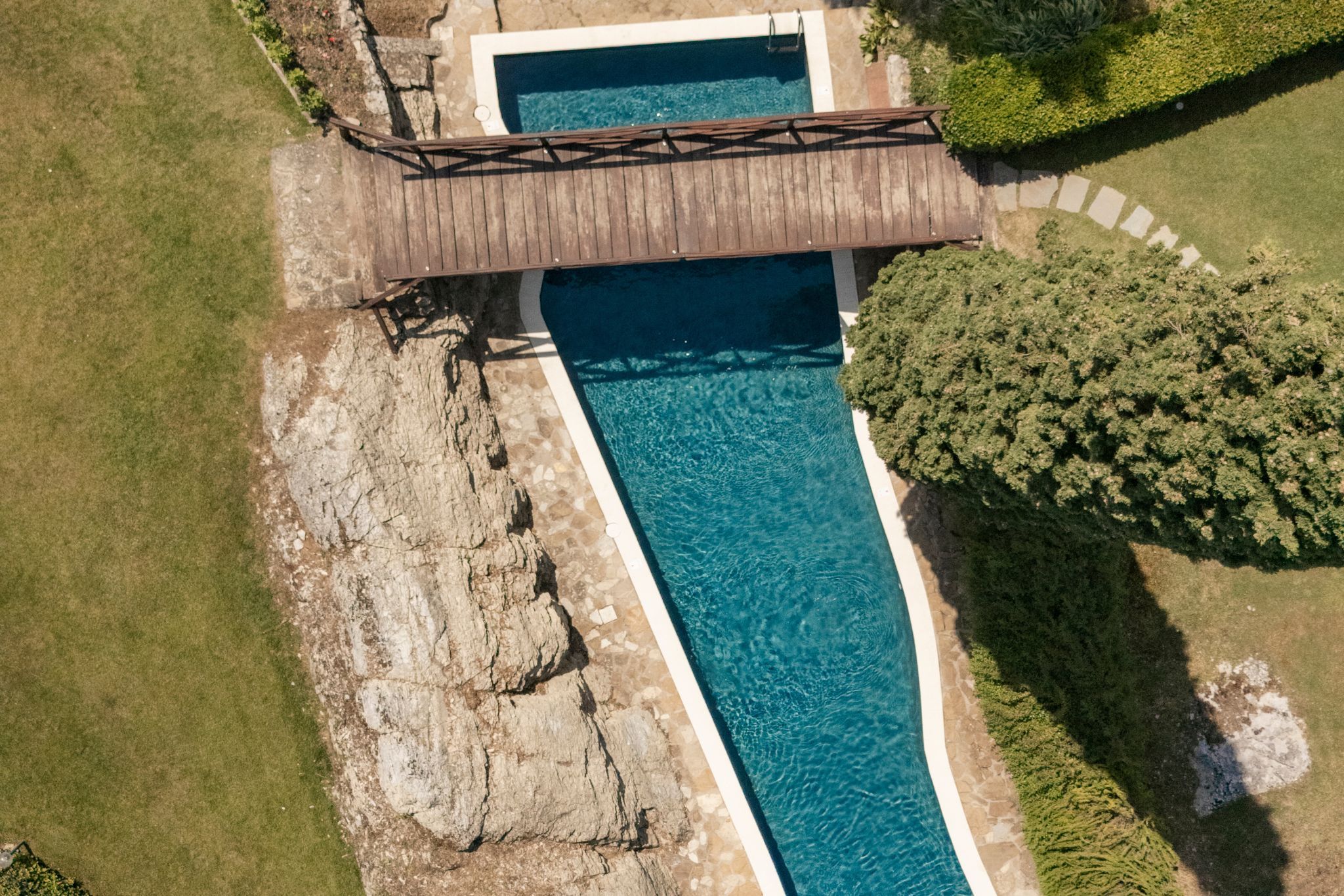 An aerial view of a swimming pool with a wooden bridge, surrounded by greenery and a rocky area.