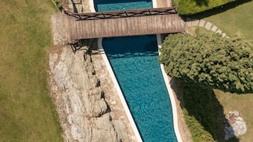 An aerial view of a swimming pool with a wooden bridge, surrounded by greenery and a rocky area.