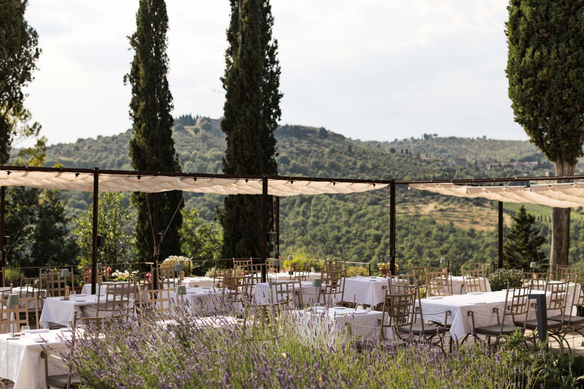 An outdoor dining area with tables and chairs, surrounded by tall trees and a view of rolling hills.