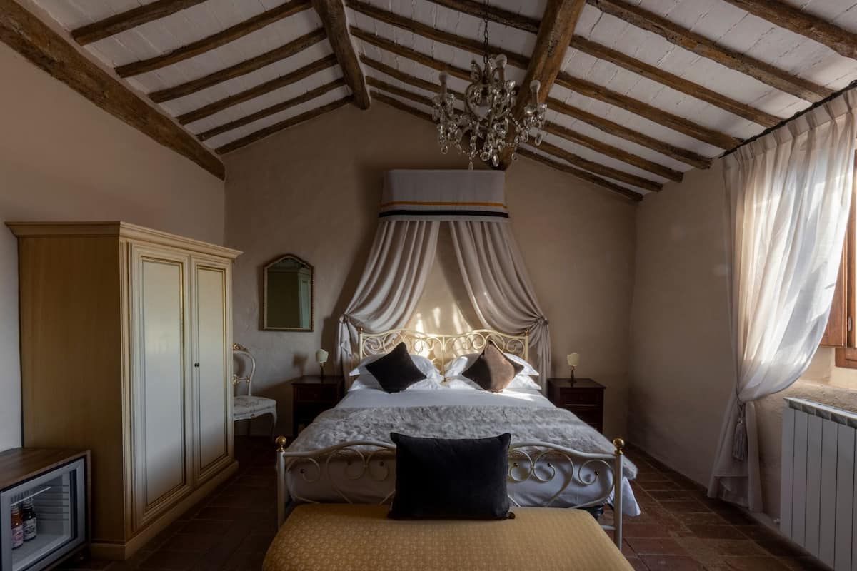 A bedroom with a canopy bed, wooden beams, a chandelier, a wardrobe, and a bedside table.