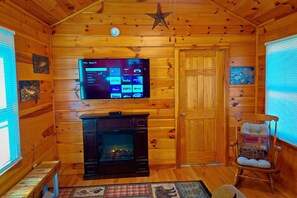 Ein gemütliches Blockhaus-Interieur mit Holzausstattung, einem an der Wand befestigten Fernseher, der Streamingdienste anzeigt, einem Kamin und einem Schaukelstuhl.