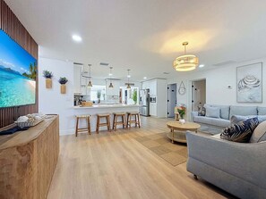 Ein modernes Wohnzimmer mit einer Couch, einem Essbereich mit Hockern, einer Küche mit weißen Schränken und einem großen Fenster mit Blick auf eine tropische Landschaft.