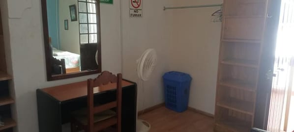 A room with a wooden desk, a chair, a mirror, a fan, a blue laundry basket, and a door with a shelf.