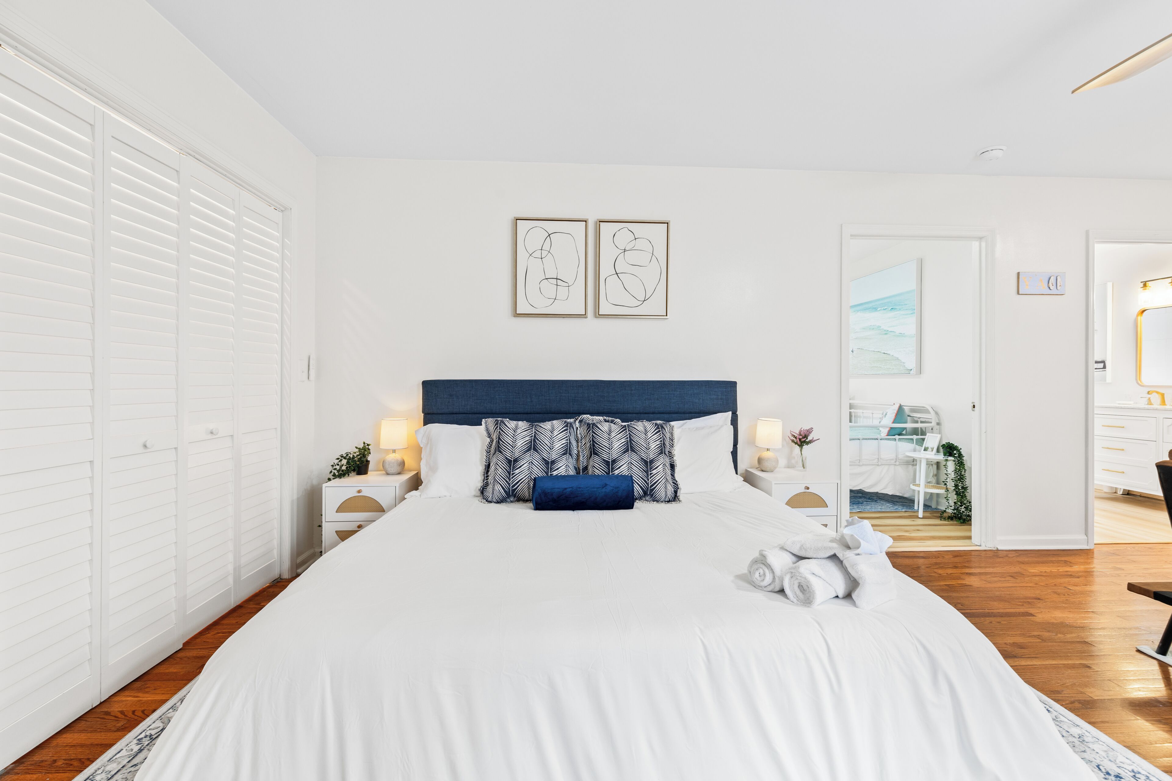 A bedroom with a large bed, white walls, a wooden floor, a headboard, two bedside tables with lamps, and a door leading to another room.