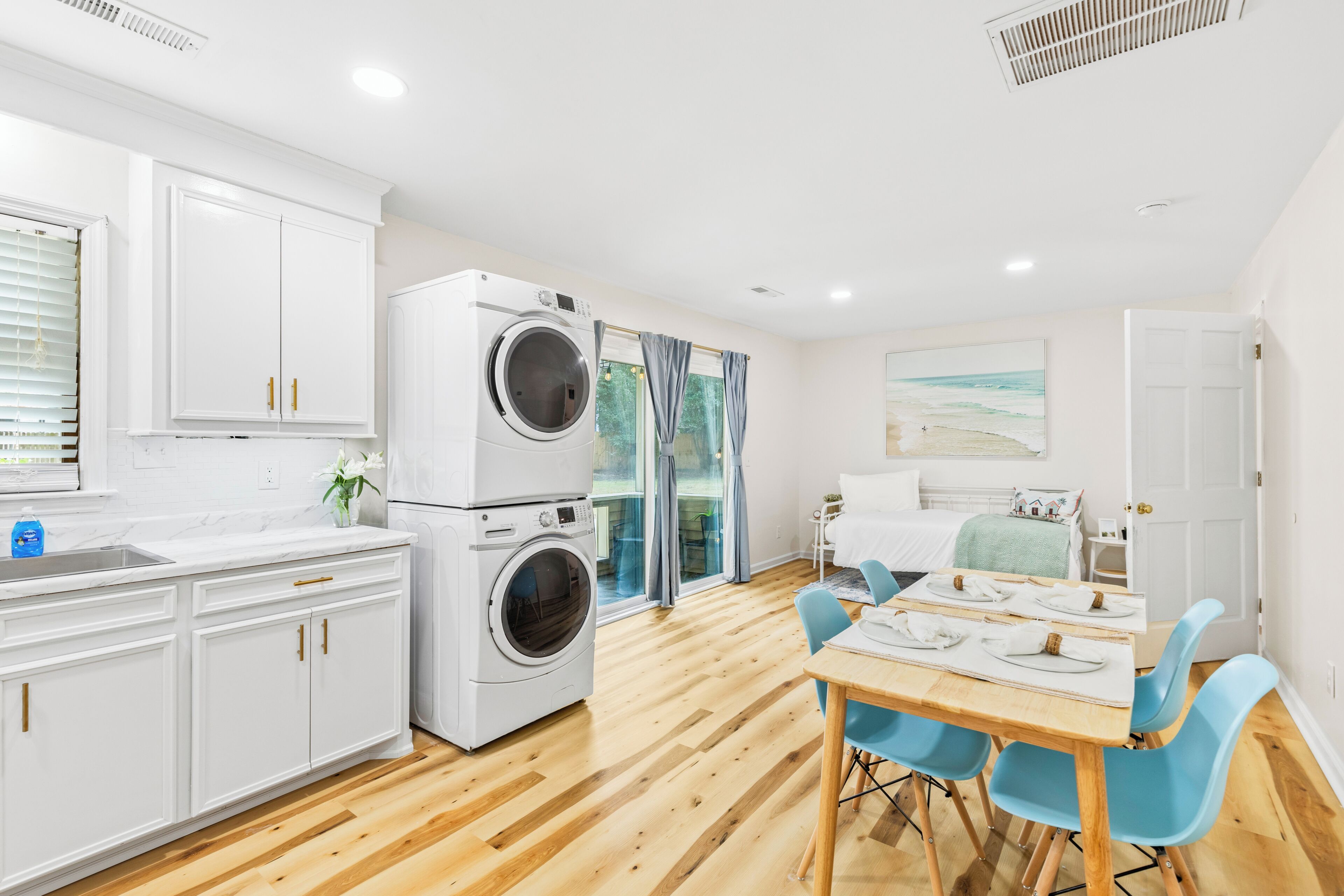 Une cuisine moderne avec des meubles blancs, un lave-linge encastré, un coin repas avec une table et des chaises, et une grande fenêtre avec des rideaux.