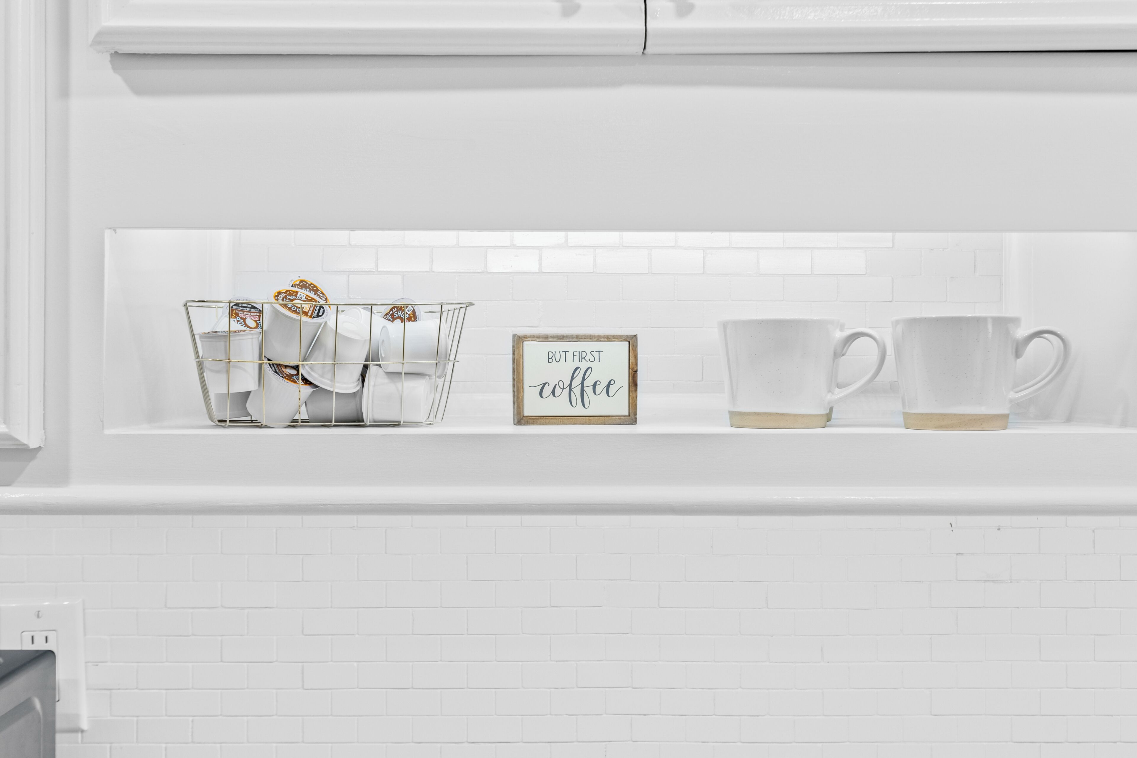 A white tiled wall with a shelf containing a glass coffee filter basket, a framed sign that says 'BUT FIRST coffee,' and two clear glass mugs.
