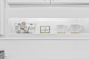 Una pared de azulejos blancos con una repisa que contiene un cesto de filtro de café de vidrio, un letrero enmarcado que dice 'BUT FIRST coffee' y dos tazas de vidrio transparentes.