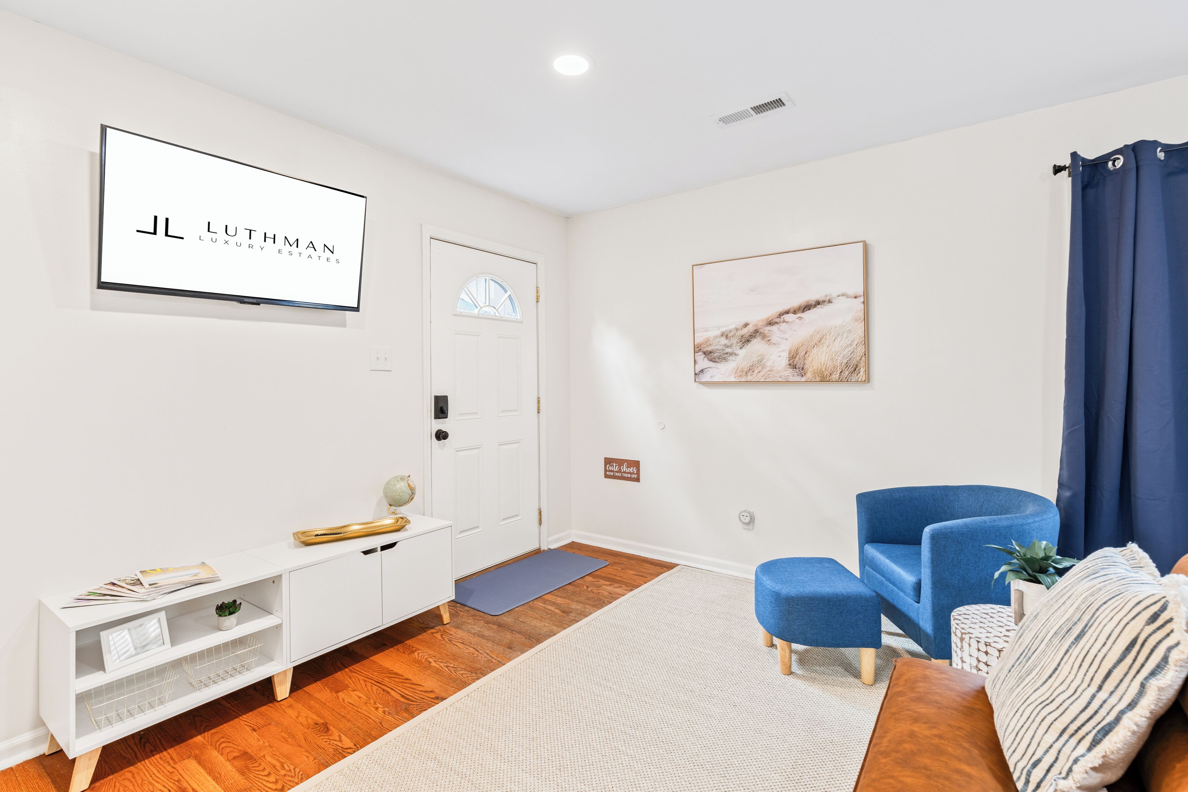 A modern living room with a white entertainment unit, a blue armchair, a sofa, and a mounted TV displaying 'JL LUTHMAN LUXURY ESTATES'.