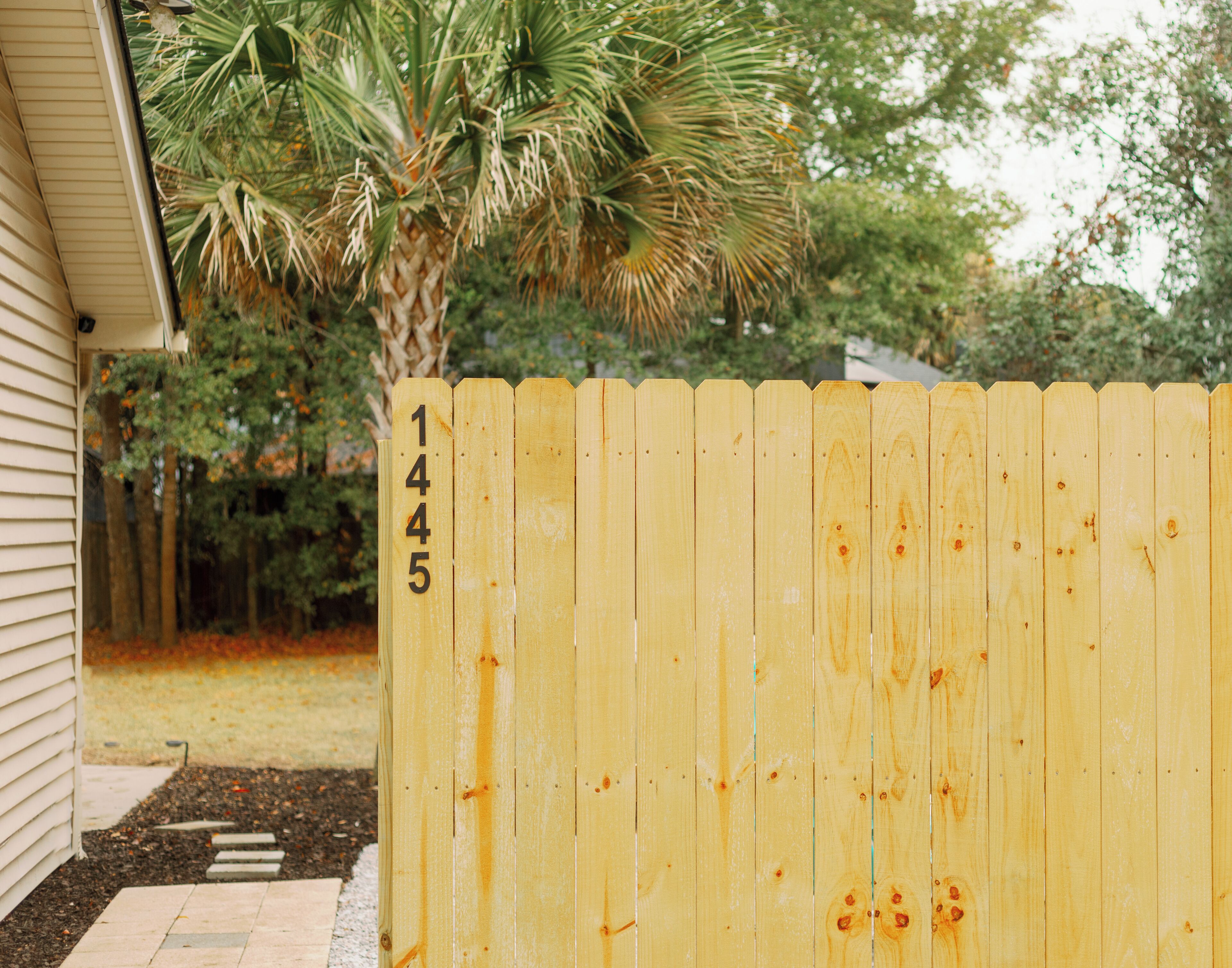 A wooden fence with the number 1445, a palm tree, and a house in the background.