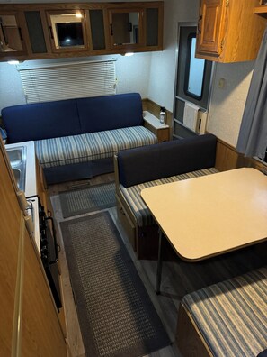 A compact RV interior with a kitchenette, a dining area, a seating area with a sofa and bench, and a small window.