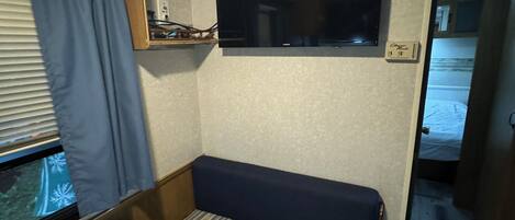 A compact RV interior with a bed, a mounted TV, and a small seating area.