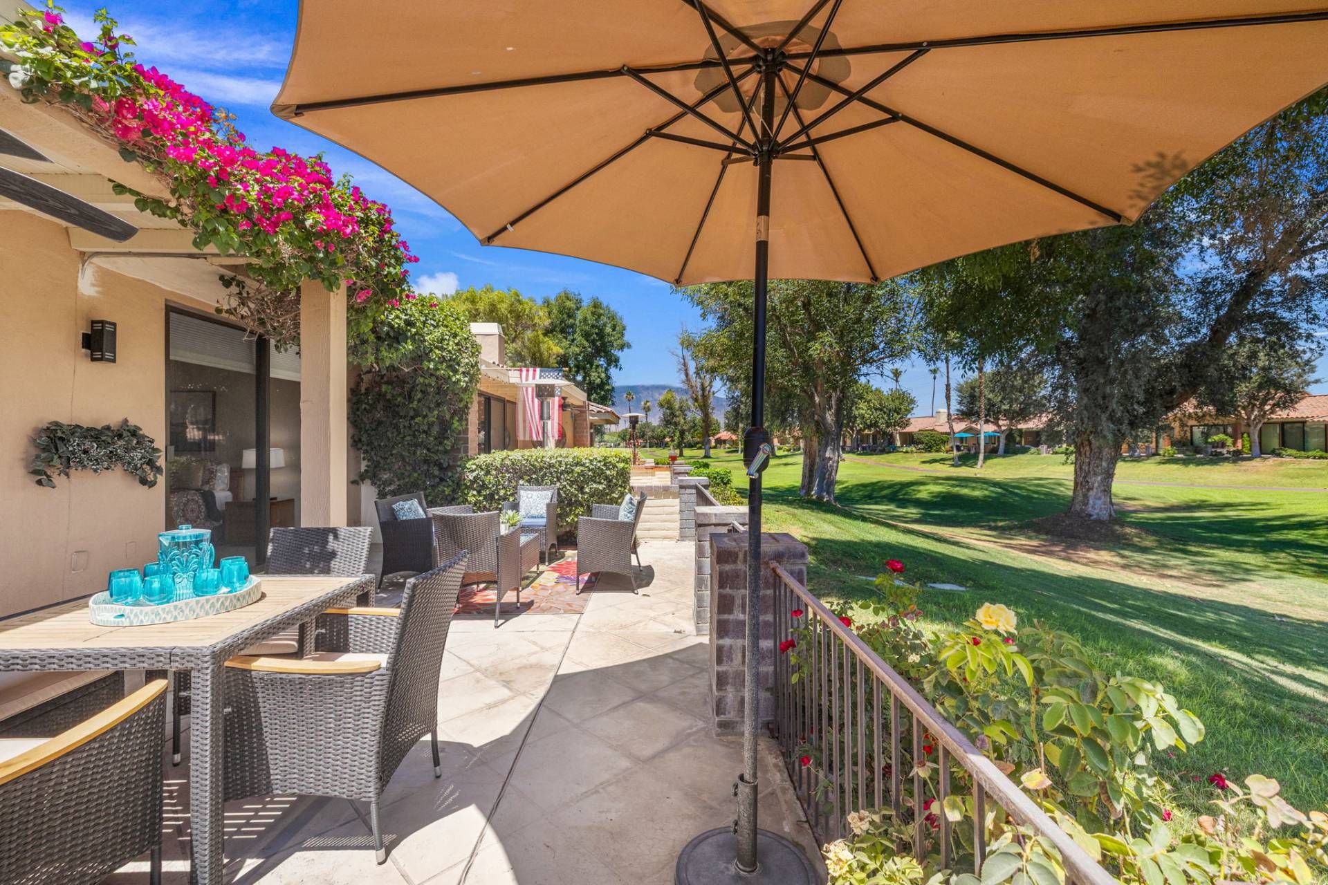 An outdoor patio with wicker furniture and a large umbrella, surrounded by greenery and a view of a golf course.