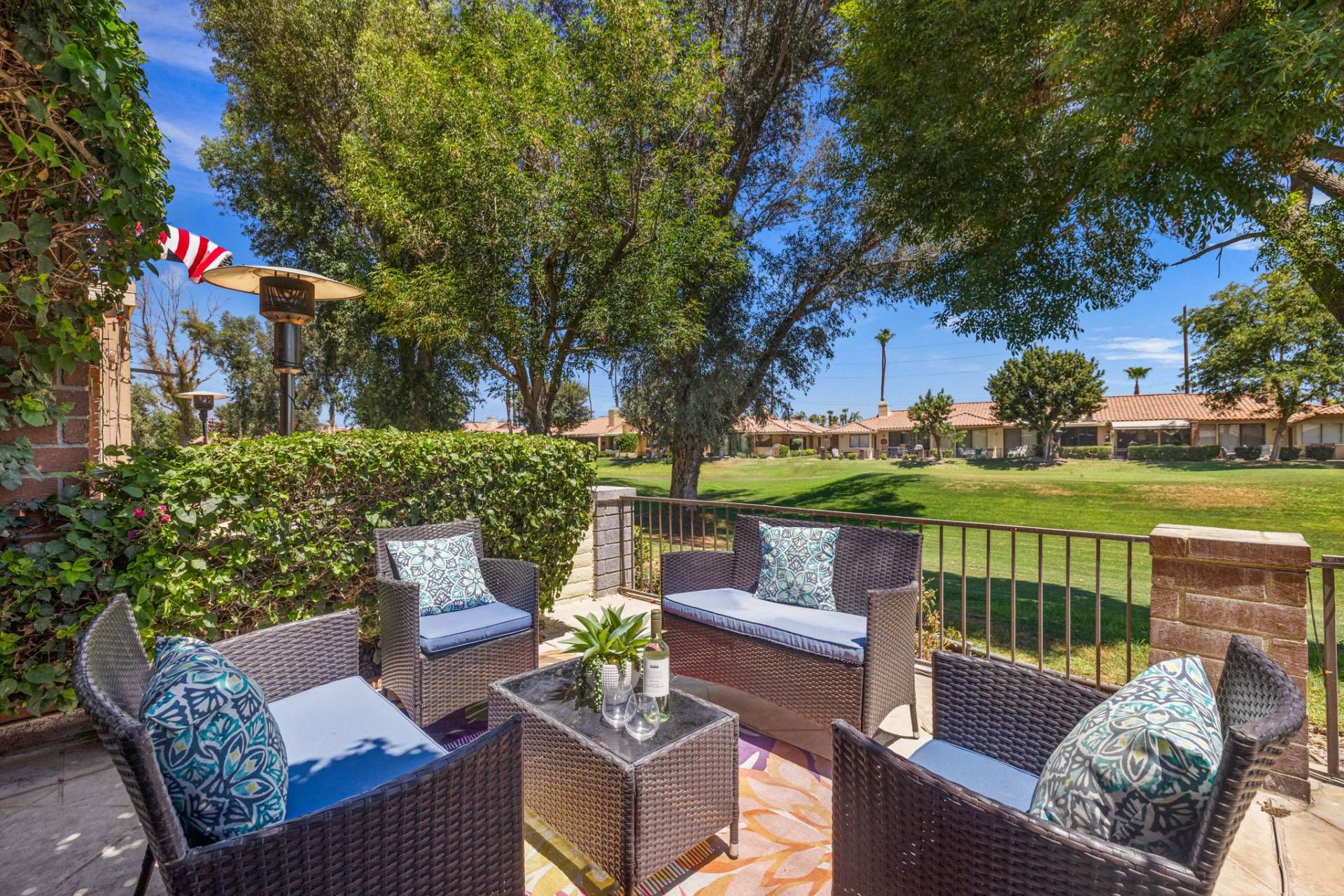 An outdoor patio with wicker furniture, a small table with a vase and wine glasses, and a view of a golf course and residential buildings in the background.