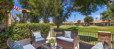 An outdoor patio with wicker furniture, a small table with a vase and wine glasses, and a view of a golf course and residential buildings in the background.