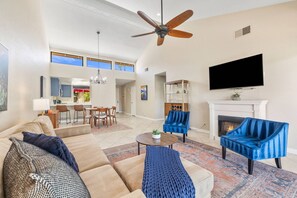 A spacious living room with a ceiling fan, a fireplace, a TV, and a dining area with a kitchen in the background. - Monterey Country Club Condo with Golf Course and Mountain Views, Pools, Spa and Private Patios (Palm Desert)