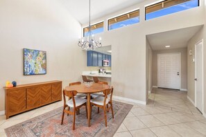 A dining area with a wooden table and chairs, a chandelier, a painting on the wall, and a view of the kitchen through an open door. - Monterey Country Club Condo with Golf Course and Mountain Views, Pools, Spa and Private Patios (Palm Desert)