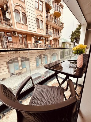 Ein Balkon mit Glastisch und Stühlen, der auf eine Straße mit Gebäuden und einem vorbeigehenden Hund blickt.