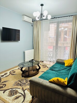 Ein Wohnzimmer mit einer Couch, einem Glastisch, einem an der Wand montierten Fernseher und einem Fenster mit Vorhängen.