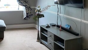Een woonkamer met een flatscreen televisie aan de muur, een grijze tv-meubel, een raam met gordijnen met een patroon, en een vloerlamp.