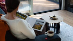 A person reading a book in a room with a small table, a cup of coffee, and a view of a wooden deck through a glass door. - Mosswood - Breath Quiet Hideaways, Hobart (Fern Tree)
