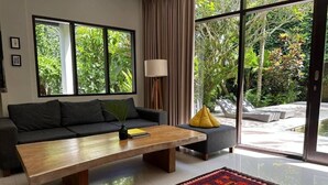 Un salón moderno con sofá, mesa de centro y vistas a un exuberante follaje a través de amplias ventanas.