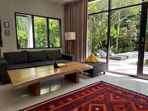 Un salón moderno con sofá, mesa de centro y vistas a un exuberante follaje a través de amplias ventanas.