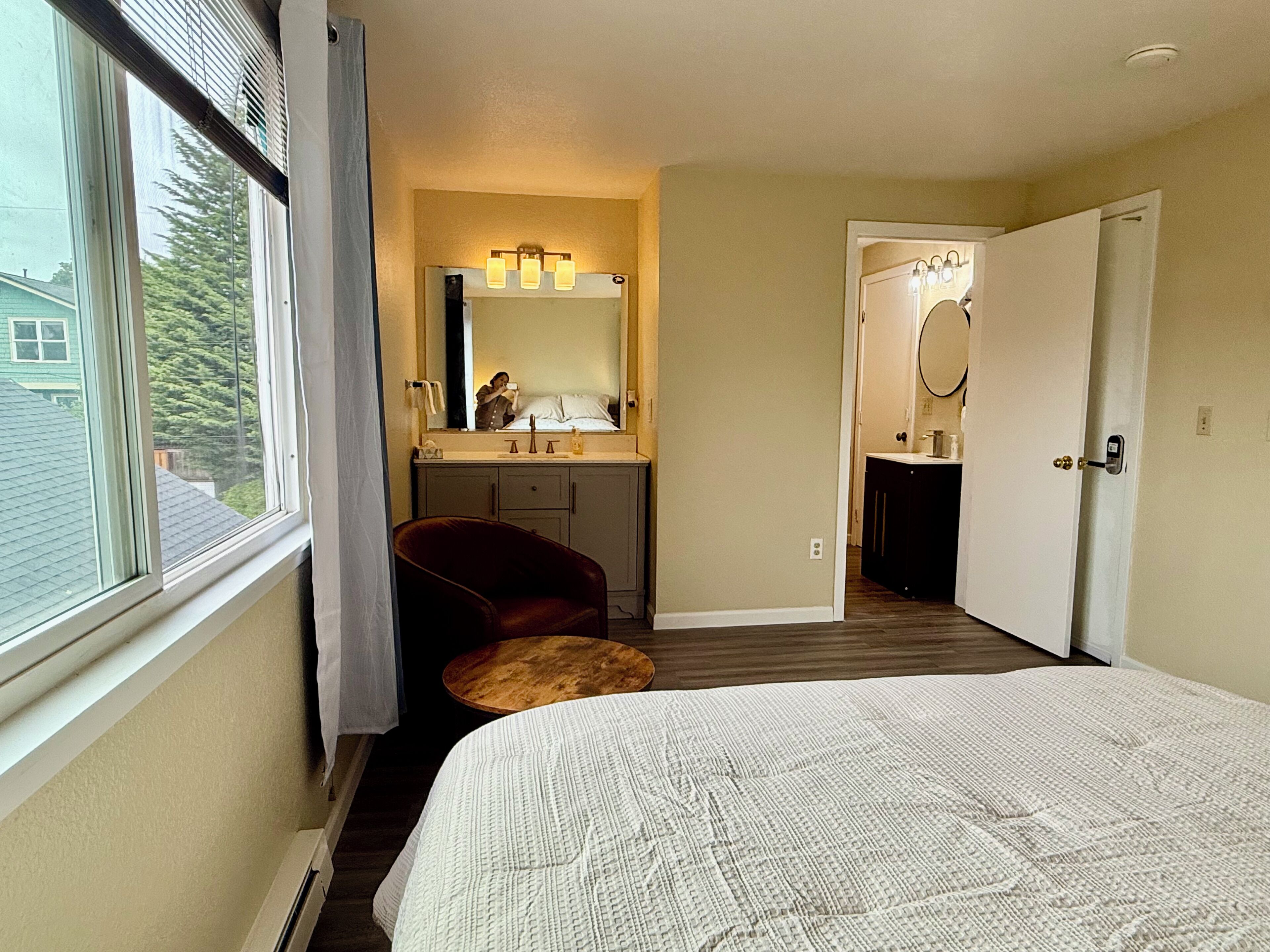 A bedroom with a bed, a chair, a vanity with a mirror, and a door leading to another room.