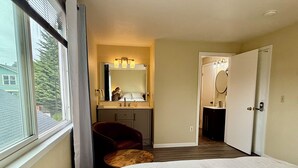 A bedroom with a bed, a chair, a vanity with a mirror, and a door leading to another room.