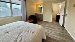 A hotel room with a bed, a chair, a small table, a bathroom with a mirror, and a door leading to another room.