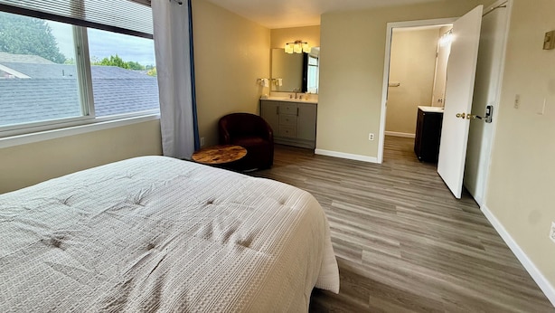 Una habitación de hotel con cama, una silla, una mesita, un baño con espejo y una puerta que da a otra habitación.