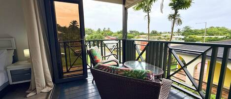 A balcony with wicker furniture, a glass table, and a view of palm trees and houses.