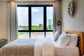 Ein Bett mit weißer Bettwäsche, ein Holz-Kopfstück und ein Blick auf Grünflächen und Gebäude durch ein Fenster mit Vorhängen.