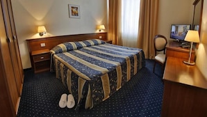 Hotellihuone, jossa on sänky, kaksi yöpöytälamppua, televisio, kirjoituspöytä lampun kanssa ja parit tohvelit lattialla.
