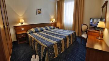 Quarto de hotel com cama, duas luminárias de cabeceira, televisão, escrivaninha com abajur e um par de chinelos no chão.