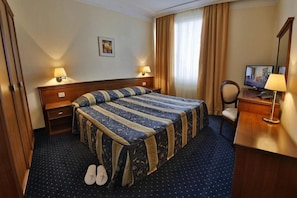 A hotel room with a bed, two bedside lamps, a television, a desk with a lamp, and a pair of slippers on the floor. - Hotel Imperial - Riviera Holiday Club Varna (Varna)