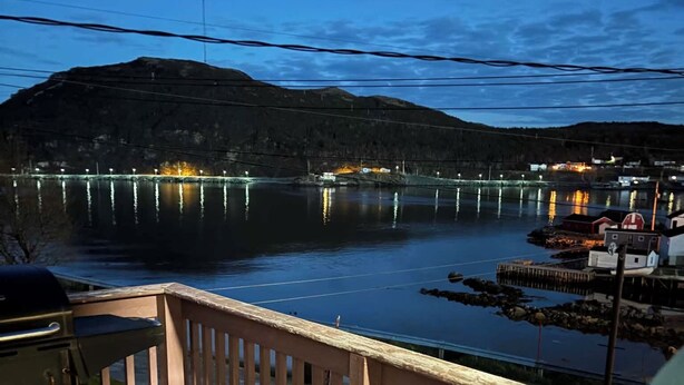 Um deck de madeira com vista para um lago calmo ao entardecer, um píer com barcos e uma linha de costa distante com edifícios.