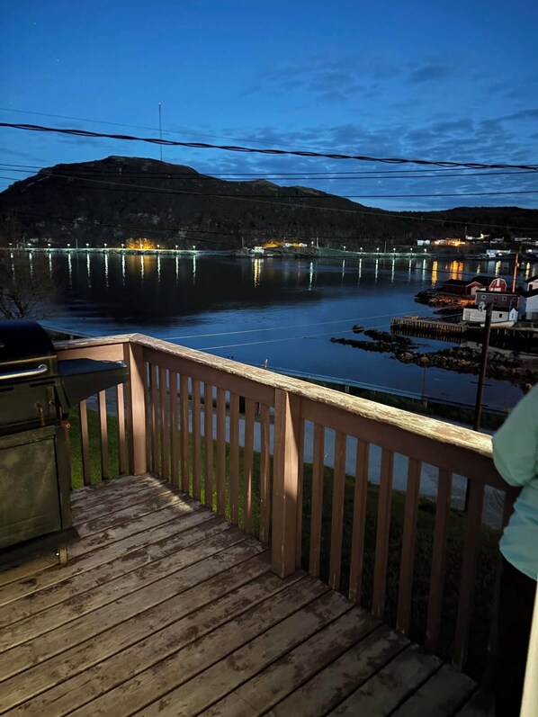 Un muelle de madera con vistas a un lago tranquilo al anochecer, un embarcadero con barcos y una orilla distante con edificios.